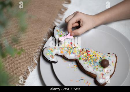 Hand des Kindes, die eine Schleife auf das Ohr des gebackenen Osterhasen-Lebkuchenkekses mit mehrfarbigen Zuckerstreuseln legt Stockfoto