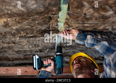 Ein Arbeiter in Overall und Schutzhelm hält einen batteriebetriebenen Bohrschraubendreher in der Hand und schraubt den Rahmen in das Holz, das er verwendet Stockfoto