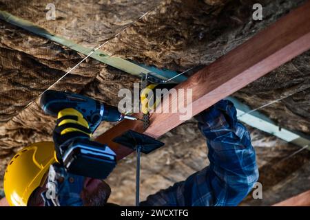 Arbeiter in Overall und Helm mit gelben Handschuhen treibt mit einem Akku-Bohrer eine Schraube in die Decke – Nahaufnahme. Stockfoto