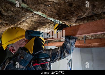 Arbeiter in Overall und Helm mit gelben Handschuhen treibt mit einem Akku-Bohrer eine Schraube in die Decke – Nahaufnahme. Stockfoto