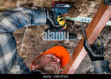 Arbeiter in Overall und Helm mit gelben Handschuhen treibt mit einem Akku-Bohrer eine Schraube in die Decke – Nahaufnahme. Stockfoto