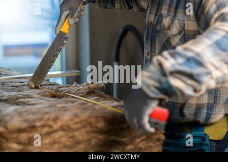 Ein Arbeiter in Overalls und Handschuhen misst die Länge der Glaswolle mit einem Maßband, das am Messer der Glaswolle befestigt ist. Stockfoto