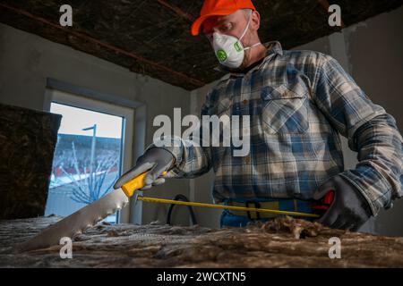 Ein Arbeiter in Overalls und Handschuhen misst die Länge der Glaswolle mit einem Maßband, das am Messer der Glaswolle befestigt ist. Stockfoto