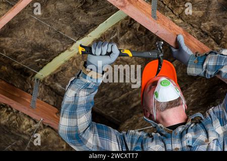 Ein Arbeiter in Overall, Handschuhen und Atemschutzmaske treibt mit einem Hammer einen Nagel ins Holz - Vorbereitung für Glaswolle im Dach. Stockfoto