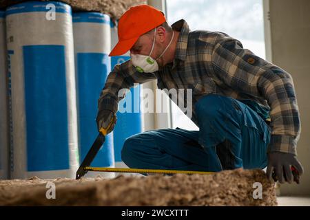 Ein Arbeiter in Overalls und Handschuhen misst die Länge der Glaswolle mit einem Maßband, das am Messer der Glaswolle befestigt ist. Stockfoto