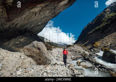 Mann mit Rucksack und Trekkingstöcken, der unter einem riesigen Felsbrocken zwischen dem Bergbach während der Wanderung zum Makalu Barun Nationalpark in Nepal spaziert. Bergwandern Stockfoto