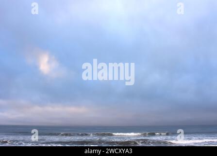 Der bewölkte Morgenhimmel wird von Blau, Violett und Rosa beleuchtet, während die Sonne an der Küste von Oregon aufgeht. Sanfte Wellen waschen an Land. Stockfoto