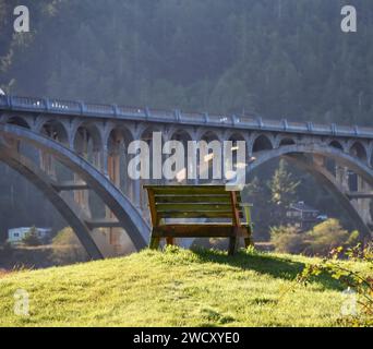 Holzbank auf Grasspitze mit Blick auf die Isaac Patterson Memorial Bridge in Oregon. Der Highway 101 überquert die Brücke und erreicht die Stadt Wedderburn. Stockfoto