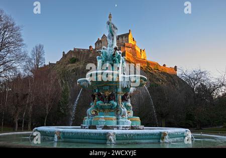 Ross Fountain, Edinburgh, Schottland, Großbritannien. 17. Januar 2024. Gefrorener Springbrunnen bei Sonnenuntergang in den West Princes Street Gardens, mit Edinburgh Castle, das in den letzten Sonnenstrahlen unter der Mondsichel leuchtet. Temperatur -1 Grad Celsius. Im Bild: Frozen Ross Fountain, der jeden Tag mehr Eiszapfen gewinnt, da die nächtlichen Temperaturen deutlich unter null Grad Celsius liegen und die Tagestemperatur bei Sonnenuntergang minus 1 Grad beträgt, sich aber wie minus 4 anfühlt. Quelle: Archwhite/Alamy Live News. Stockfoto