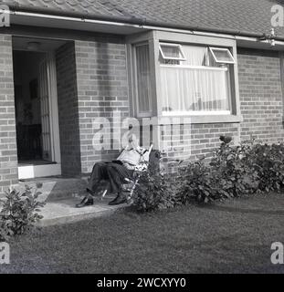 1960er Jahre, historisch, ein Gentleman, der in einem Liegestuhl sitzt und draußen ein Buch liest, auf der ersten Stufe eines neuen Chalet-Bungalows in England, Großbritannien. Stockfoto