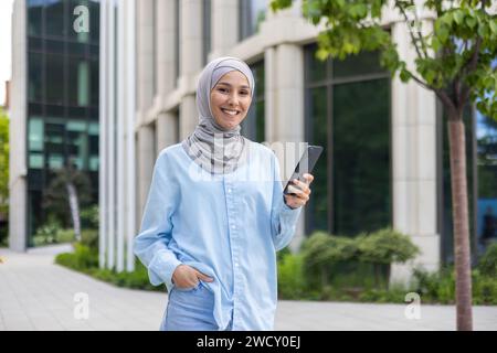 Eine junge, fröhliche muslimische Frau, die einen Hijab trägt und ein Smartphone hält, geht selbstbewusst an einem Bürogebäude vorbei. Stockfoto