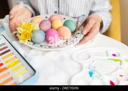 Ein Mädchen, das einen Teller mit bunt bemalten Ostereiern mit einer Farbpalette und einem Pinsel in der Nähe hält. Die Eier sind in Pastelltönen wie Blau, pi bemalt Stockfoto
