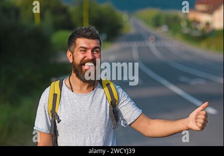 Reisen mit Autostop, mit Sommerausflug. Auto-Stopp-Fahrt. Mann mit strengem Gesicht und Bart, der durch Anhalter mit Straße im Hintergrund reist. Stockfoto