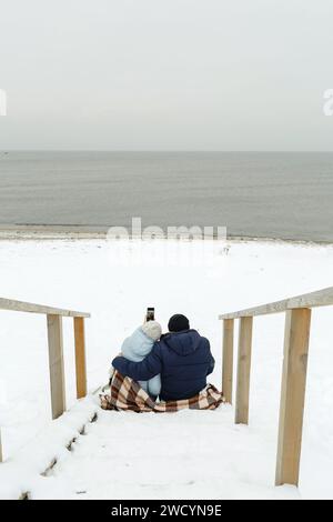 Glückliches älteres Paar, das im Winter auf der Treppe am Meer sitzt und ein Selfie macht. Vertikaler Rahmen. Stockfoto