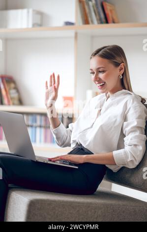 Frau, die Videoanrufe auf dem Laptop hat, während sie in ihrer flachen, winkenden Hand sitzt und Hallo winkt. Stockfoto