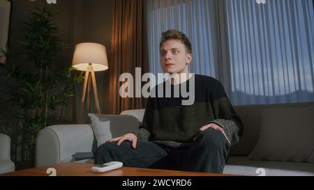 Der blonde junge Mann sitzt abends zu Hause auf der Couch und sieht sich einen Film im Fernsehen an Stockfoto