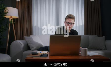 Junge blonde Haarstudentin mit Brille, die von zu Hause aus mit Laptop arbeitet, während sie zu Hause auf dem Sofa sitzt. Fokussierter Millennial-Mann, der auf l Stockfoto
