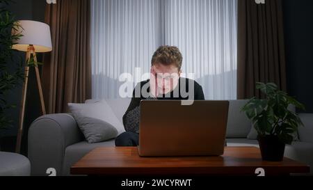Junger blonder Mann, der zu Hause auf dem Sofa sitzt, während er einen Laptop benutzt, der Angst und Stress hat. Verärgerter junger Mann, der auf Verlust reagiert, schlechte Nachrichten auf dem Laptop Stockfoto