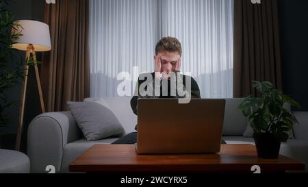 Hübscher blonder Mann im Teenageralter sitzt zu Hause auf dem Sofa, während er einen Laptop benutzt und Angst und Stress hat. Verärgerter junger Mann, der auf Verlust reagiert, schlechte Nachrichten auf dem Laptop Stockfoto