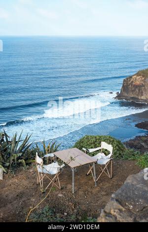 Eine ruhige Szene mit zwei leeren Campingstühlen und einem Tisch, einladend auf einer Klippe mit Blick auf das Meer aufgestellt, bei hellem Tageslicht Stockfoto