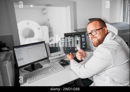 Medizinische Computertomographie oder MRT-Scanner. Arzt sitzt am Computer. Männlicher Spezialist mit Brille und weißem Gewand, Blick in die Kamera, Lächeln, Daumen hoch zeigen. Konzept der modernen Diagnostik. Stockfoto