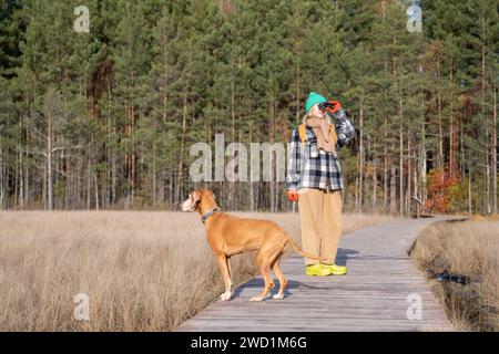 Frau mit Jagdhund, der auf ökologischem Weg im Herbstwald durch ein Fernglas schaut. Stockfoto