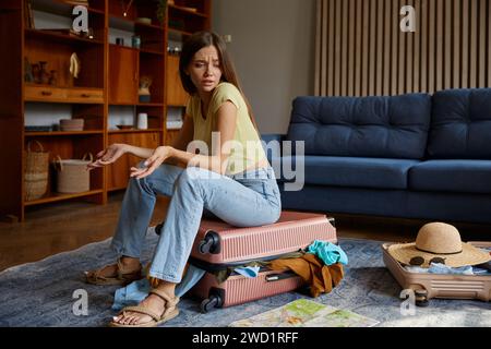 Frustrierte junge Frau, die auf Reisekoffer sitzt und Kleidung überzogen hat Stockfoto