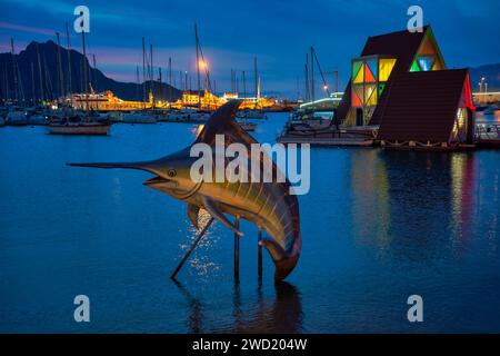 Dämmerungsblick auf einen Yachthafen in Mindelo, Kap Verde, mit einer metallischen Skulptur eines Marlins im Vordergrund mit beleuchteten dreieckigen Gebäuden A Stockfoto