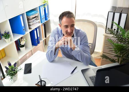 Freischaffender Unternehmer lächelnd, der in seinem Heimbüro einen Laptop ansieht und stolz auf einen Laptop in einem weißen Büro mit Regalen Fu auf einem Stuhl sitzt Stockfoto