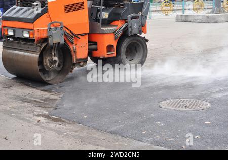 Die Straßenwalze oder Dampfwalze verdichtet sich und pflastet heißer Asphalt um das Mannloch auf dem Asphaltbelag. Stockfoto