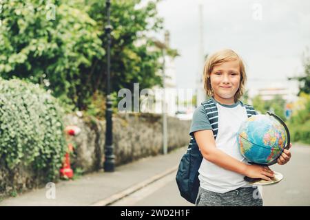 Außenporträt eines lustigen kleinen Jungen mit Rucksack und Weltkugel. Back-to-School-Konzept. Filterbild für Film-Look Stockfoto