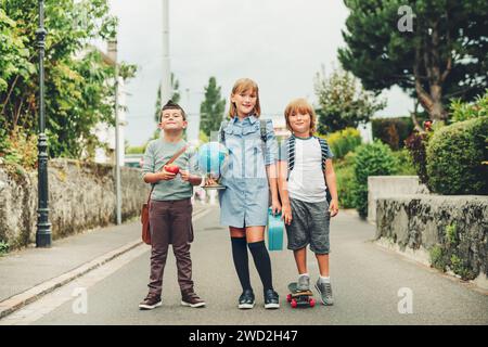 Eine Gruppe von drei lustigen Kindern, die Rucksäcke tragen und zurück zur Schule gehen. Mädchen und Jungen genießen Schulaktivitäten. Globus, Lunchbox, roter Apfel und Skateboa Stockfoto