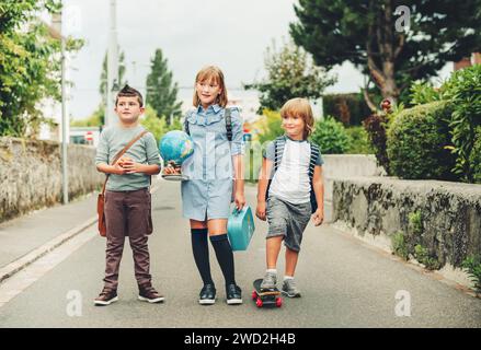 Eine Gruppe von drei lustigen Kindern, die Rucksäcke tragen und zurück zur Schule gehen. Mädchen und Jungen genießen Schulaktivitäten. Globus, Lunchbox, roter Apfel und Skateboa Stockfoto