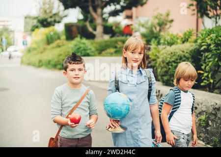 Eine Gruppe von drei lustigen Kindern, die Rucksäcke tragen und zurück zur Schule gehen. Mädchen und Jungen genießen Schulaktivitäten. Globus, Lunchbox, roter Apfel und Beutel Stockfoto