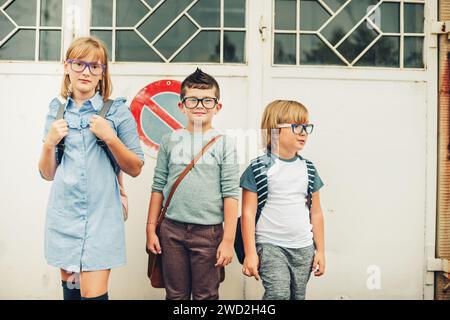 Eine Gruppe von drei lustigen Kindern, die Rucksäcke tragen und zurück zur Schule gehen. Mädchen und Jungs mit Brille, die draußen posieren Stockfoto