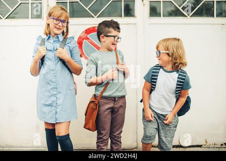 Eine Gruppe von drei lustigen Kindern, die Rucksäcke tragen und zurück zur Schule gehen. Mädchen und Jungs mit Brille, die draußen posieren Stockfoto