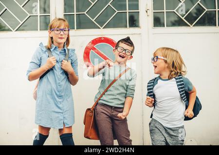 Eine Gruppe von drei lustigen Kindern, die Rucksäcke tragen und zurück zur Schule gehen. Mädchen und Jungs mit Brille, die draußen posieren Stockfoto