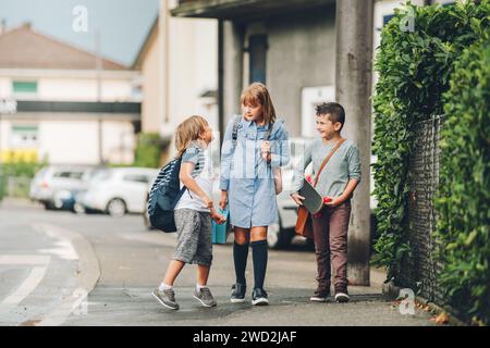 Gruppe von 3 lustigen Schulkindern, die gemeinsam zurück zur Schule gehen, Rucksäcke tragen, Lunchbox halten und Skateboard fahren Stockfoto