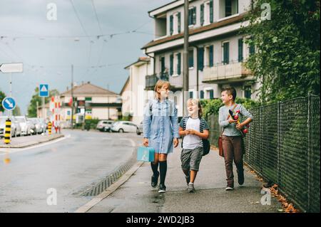 Gruppe von 3 lustigen Schulkindern, die gemeinsam zurück zur Schule gehen, Rucksäcke tragen, Lunchbox halten und Skateboard fahren Stockfoto