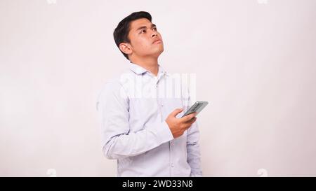 Erwachsener asiatischer Mann, der an etwas denkt, während er das Handy vor weißem Hintergrund hält Stockfoto