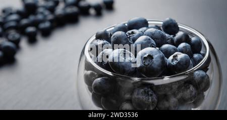 Nahaufnahme eines Glastopfes voller Erdbeeren auf schwarzem Stein. Ein Haufen verstreuter Heidelbeeren im Hintergrund. Selektiver Fokus. Stockfoto