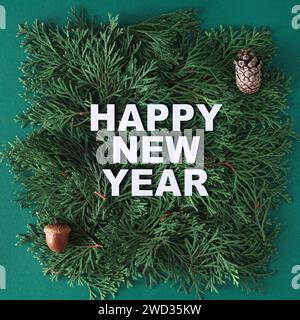 Happy New Year Text aus weißem Papier auf Thuja-Zweigen mit Eichel und Kiefernkegel auf grünem Hintergrund. Kreatives Neujahrskonzept. Stockfoto