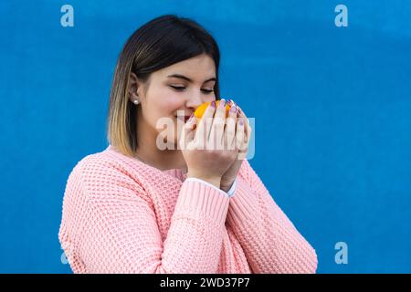 Eine lächelnde junge kurvige Frau in einem rosafarbenen Pullover genießt das Aroma einer frischen Orange vor einem hellblauen Hintergrund und ruft Frische und Wohlbefinden hervor. Stockfoto