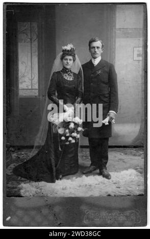 Studio-Porträt von Jungvermählten in voller Länge. Die Braut hat einen Schleier auf dem Kopf und hält einen Strauß weißer Rosen in der Hand. Der Bräutigam trägt Stockfoto