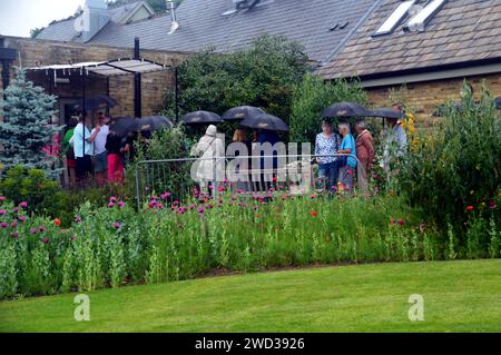 Die Leute standen in der Warteschlange im Regen und hielten Regenschirme vor Betty's Tea Room im RHS Garden Harlow Carr, Harrogate, Yorkshire, England Stockfoto