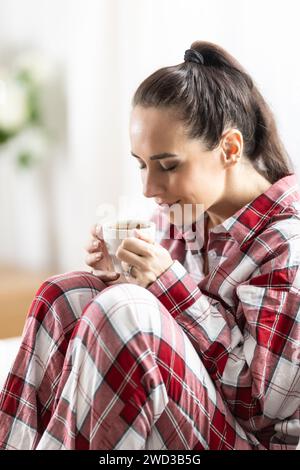 Junge Frau im rot karierten Pyjama, die auf dem Sofa sitzt und morgens Kaffee im Wohnzimmer trinkt. Stockfoto