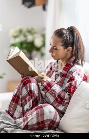 Die schöne Brünette im Pyjama liest ein Buch, das auf dem Sofa im Wohnzimmer sitzt. Stockfoto