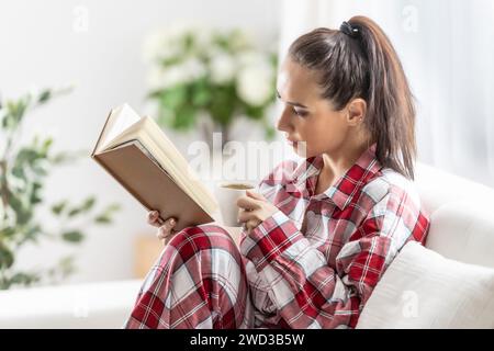 Die schöne Brünette im Pyjama liest ein Buch, das auf dem Sofa im Wohnzimmer sitzt. Stockfoto