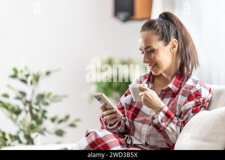 Am Morgen findet eine junge Brünette im Pyjama heraus, was andere in sozialen Netzwerken Neues haben, trinkt Kaffee und sitzt auf dem Sofa im Wohnzimmer. Stockfoto