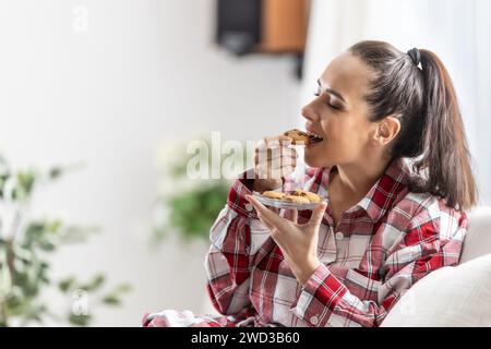 Eine junge Frau, die noch im Pyjama sitzt, genießt süße Schokoladenkekse auf dem Sofa im Wohnzimmer. Stockfoto